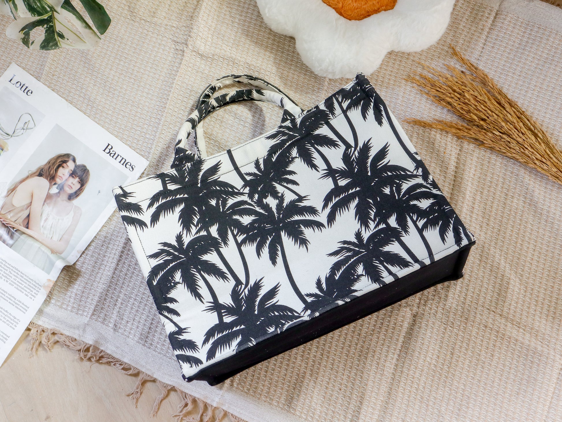 Tote bag with palm tree pattern on a textured surface with magazines and flowers.